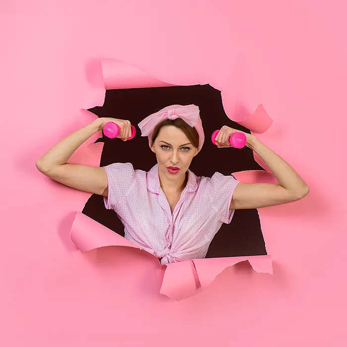 Egy nő erőt és magabiztosságot sugároz, miközben rózsaszín háttérből tör ki, mindkét kezében súlyzóval, ami a kreatív és erőteljes márkaépítés erejét jelképezi.